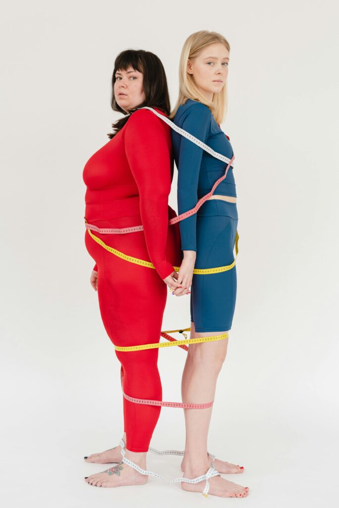 Full body of barefoot plus size woman with slender female in activewear covered with yellow measuring tapes looking at camera on white background